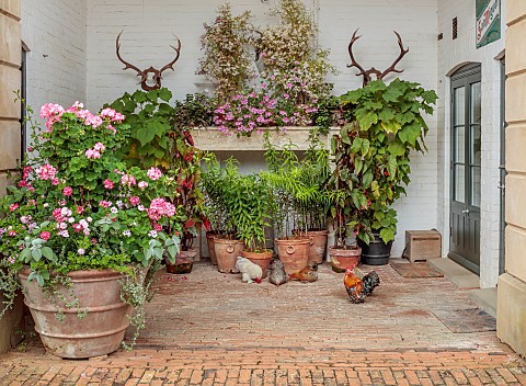 ADMINGTON_HALL_WARWICKSHIRE_COURTYARD_TERRACOTTA_CONTAINERS_FILLED_WITH_LILLIES_GERANIUMS_FIREPLACE_