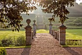 EASTON WALLED GARDENS, LINCOLNSHIRE: STEPS DOWN TO LAWN WITH CLIPPED TOPIARY CONES. BRIDGE OVER THE RIVER WITHAM. COUNTRY GARDEN, ENGLISH GARDEN, FORMAL, SUMMER, DAWN, SUNRISE