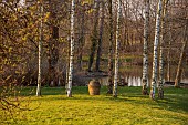 PRIORS MARSTON, WARWICKSHIRE, THE MANOR HOUSE: MARCH, TERRACOTTA CONTAINER SURROUNDED BY CIRCLE OF WHITE BIRCHES, LAKE