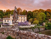 IFORD MANOR, WILTSHIRE: MAY, SPRING, PURPLE FLOWERS OF WISTERIA SINENSIS ON THE MANOR HOUSE SEEN FROM THE BRIDGE OVER THE RIVER FROME, EVENING LIGHT