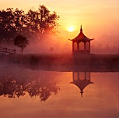 DAWN MIST: SUN RISES OVER CHINESE STYLE PAGODA BESIDE LAKE IN PRIVATE GARDEN.  CHASTLETON GLEBE OXFORDSHIRE
