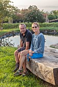 NEW WOOD TREES, DEVON: TIFFANY AND PHILIP NIEUWOUDT SITTING ON A HUGE WOODEN BENCH SCULPTURE BY POND