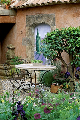 TABLE__CHAIRS_ON_COBBLE_TERRACE_WITH_TROMPE_LOEIL_PAINTING_BY_JOHN_SIMPSON_IN_BG_BSKYB_GARDEN_DESIGN