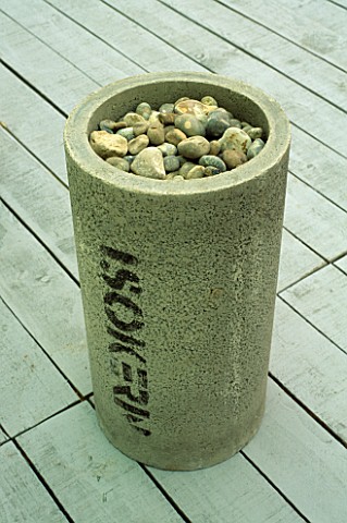 SIMPLE_SCULPTURAL_FORM_OF_CONCRETE_CYLINDER_FILLED_WITH_STONES_STANDS_ON_WOODEN__DECKING_IN_MODERN_R
