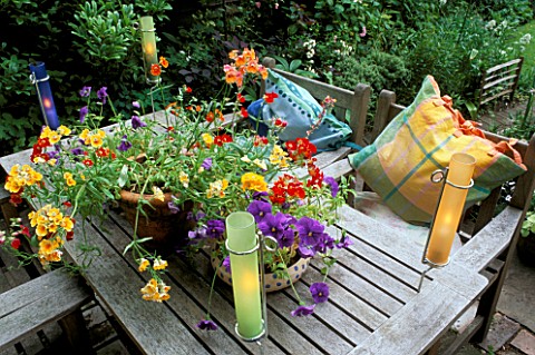 CANDLES_IN_COLOURED_GLASS_TUBES_LIGHT_UP__A_GROUP_OF_SUMMER_CONTAINERS_ON_TABLE_SEATS_WITH_CUSHIONS_
