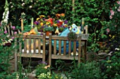 SHELTERED CORNER OF GARDEN WITH TABLE & CHAIRS. CANDLES IN COLOURED GLASS TUBES LIGHT UP COLOURFUL CUSHIONS & FLORAL ARRANGEMENT ON TABLE. DESIGNER: LISETTE PLEASANCE