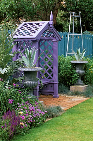 BLUE_FENCE__MAUVE_COVERED_WOODEN_SEAT__PEWTERED_URNS_PLANTED_WITH_AGAVE_AMERICANA_AND_CARDOONS_THE_N