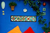DECORATIVE FEATURE: BRIGHTLY COLOURED MOSAIC SET IN ULTRAMARINE BLUE WALL. BENEATH ON A WOODEN BENCH ARE ORANGE AND RED CUSHIONS. DESIGNERS: ANDREW AND KARLA NEWELL