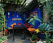 CENTRAL COURTYARD PARTIALLY COVERED BY A TIMBER PERGOLA. TABLE AND BENCHES WITH ORANGE AND RED CUSHIONS BESIDE ULTRAMARINE WALLS DECORATED WITH MOSAIC PANELS AND MEXICAN TILES.