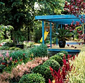 BLUE PAINTED PERGOLA OVER DECKING WITH BOX BALLS AND ASTILBES IN THE FOREGROUND. TO THE BACK IS A YELLOW CHILDRENS SLIDE. DESIGN BY DAVID STEVENS