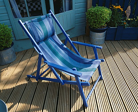 A_PLACE_TO_SIT_BLUE_STRIPED_CANVAS_DECKCHAIR_ON_RIBBED_DECKING_IN_BG_IS_PAINTED_SHED_AND_METAL_CONTA