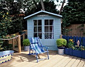 PAINTED BEACH HUT STYLE SHED STANDS BEHIND BLUE STRIPED CANVAS DECKCHAIR  PAINTED TIN BATH AND RIBBED DECKING. ROBIN GREEN AND RALPH CADES SEASIDE STYLE GARDEN  LONDON.