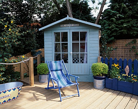 PAINTED_BEACH_HUT_STYLE_SHED_STANDS_BEHIND_BLUE_STRIPED_CANVAS_DECKCHAIR__PAINTED_TIN_BATH_AND_RIBBE