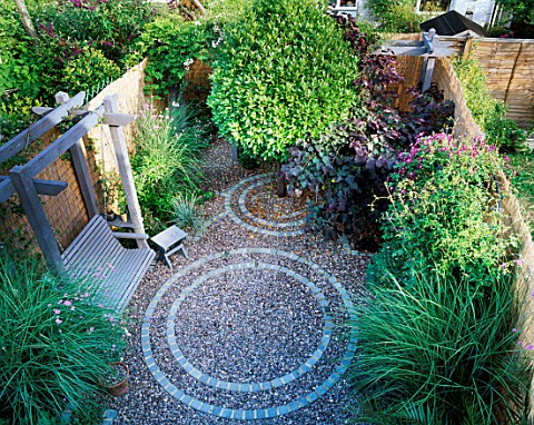 SMALL_GARDEN_WITH_CIRCULAR_BRICK_PATTENS_IN_GRAVEL_WITH_NATURAL_WOODEN_SWING_SEAT_AND_STOOL__VERBENA