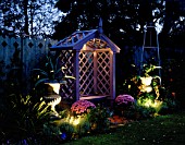 NICHOLS GARDEN: COVERED WOODEN SEAT SURROUNDED BY TWO SILVER PAINTED METAL URNS PLANTED WITH AGAVES. IN TWO POTS ARE PINK CHRYSANTHEMUMS. LIGHTING BY GARDEN & SECURITY LIGHTING