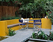 A PLACE TO SIT: ALUMINIUM TABLE AND CHAIRS ON PATIO SURROUNDED BY YELLOW RENDERED WALLS WITH RAISED BEDS AND RILL. TRACHYCARPUS AND PHORMIUM FOLIAGE IN B/G . DESIGNER JOE SWIFT