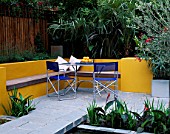 A PLACE TO SIT: ALUMINIUM TABLE AND CHAIRS ON PATIO SURROUNDED BY YELLOW RENDERED WALLS WITH RAISED BEDS AND OLEANDER   TRACHYCARPUS. DESIGNER JOE SWIFT