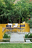 A PLACE TO SIT: ALUMINIUM TABLE AND CHAIRS ON PATIO SURROUNDED BY YELLOW RENDERED WALLS AND  RILL WITH OLEANDER   TRACHYCARPUS. DESIGNER JOE SWIFT