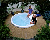 HAZEL AND ROBERT PLAY IN THE SPA POOL SURROUNDED BY PALMS IN POTS AND DECKING. HAMPTON COURT 2000  DESIGNER BOARDMAN GELLY & CO