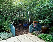 CHILDRENS SWINGS WITH BARK CHIP UNDERNEATH  BLUE TRELLIS  FIG (FICUS BROWN TURKEY)   FATSIA JAPONICA AND TERRACOTTA WATER FEATURE.