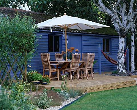 BLUE_SUMMERHOUSE_WITH_DECKING__TABLE__CHAIRS_AND_PARASOL__BIRCH_TREE_TRUNKS__ORNAGE_LILLIES__HAMMOCK