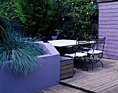 GARDEN WITH DECKING DESIGNED BY JOE SWIFT AND THAMASIN MARSH:  LILAC/GREY SHED AND RENDERED WALL OF RAISED BED WITH BLUE FESTUCAS  STONE TOPPED TABLE AND CHAIRS