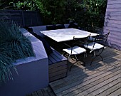 GARDEN WITH DECKING. DESIGN JOE SWIFT AND THAMASIN MARSH: LILAC/GREY RAISED BED WITH RENDERED WALL AND BLUE GRASSES.  STONE TOPPED TABLE  CHAIRS AND BENCHING WITH CUSHIONS.   SHED