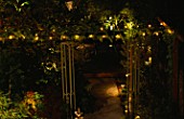 OVERVIEW OF GARDEN AT NIGHT  DESIGNED BY JOE SWIFT WITH OBELISKS LIT BY GARDEN & SECURITY LIGHTING.  FAIRY LIGHTS RUN ACROSS GARDEN ON WIRES.