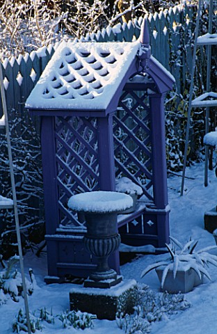 PURPLE_COVERED_SEAT_FLANKED_BY_METAL_URNS_AND_BACKED_BY_BLUE_DECORATIVE_FENCING_IN_THE_NICHOLS_GARDE