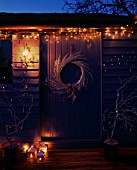 THE BLUE SUMMERHOUSE AT NIGHT  WITH SILVER WREATHE  TWISTED WILLOW TREES IN PURPLE PLASTIC CONTAINER  CANDLE AND FAIRY LIGHTS