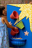 NANCY PLAYS IN THE BUCKET WATER CASCADE: BLUE BOARD WITH YELLOW MOSAIC  COLOURED TERRACOTTA POTS  SHELLS  STARFISH AND RUNNING WATER