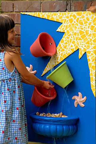 NANCY_PLAYS_IN_THE_BUCKET_WATER_CASCADE_BLUE_BOARD_WITH_YELLOW_MOSAIC__COLOURED_TERRACOTTA_POTS__SHE