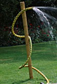 WATER FEATURE: WATER SERPENT MADE FROM YELLOW HOSE TIED WITH TWINE TO BAMBOO POLE IN LAWN. DESIGNER: CLARE MATTHEWS