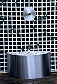 WATER FEATURE: STAINLESS STEEL DISC AND SPOUT POURS INTO A STAINLESS STEEL CONTAINER WITH WHITE PEBBLES. WHITE TRELLIS BEHIND. DESIGN: CLARE MATTHEWS. TRELLIS BY ASTOR TRELLISWORK