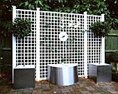 WATER FEATURE: STAINLESS STEEL DISC AND SPOUT POURS INTO CONTAINER WITH WHITE PEBBLES. WHITE TRELLIS BEHIND. DESIGN: CLARE MATTHEWS. TRELLIS BY ASTOR TRELLISWORK