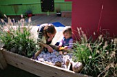 CHILDRENS DECK GARDEN: LUCY  AND CLARE FEELING THE SHELLS  FIR CONES AND STONES IN A RAISED WOODEN BED: DESIGNER: CLARE MATTHEWS