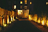 TOWN GARDEN LIT UP AT NIGHT WITH BRICK WALL  OLIVE TREES  LAWN AND HOUSE IN THE BACKGROUND. DESIGNER: CLAIRE MEE