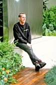 GARDEN DESIGNER STEPHEN WOODHAMS SITS ON THE WATER FEATURE. SANCTUARY GARDEN SPONSORED BY MERRILL LYNCH AT THE CHELSEA FLOWER SHOW 2002: DESIGNED BY STEPHEN WOODHAMS: