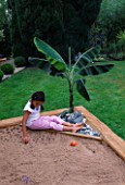 DESIGNER: CLARE MATTHEWS - TROPICAL ISLAND SANDPIT - WOODEN FRAMEWORK WITH CROSS FRAME AND PLASTIC SHEETING  BANANA  PEBBLES  SAND AND GIRL