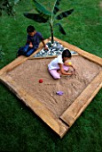 DESIGNER: CLARE MATTHEWS - TROPICAL ISLAND SANDPIT - WOODEN FRAMEWORK WITH CROSS FRAME AND PLASTIC SHEETING  BANANA  PEBBLES  SAND  GIRL AND BOY