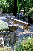 LA CHABAUDE  FRANCE. DESIGNER - PHILIPPE COTTET: A PLACE TO SIT - GRAVEL TERRACE WITH LONG WOODEN BENCH PROPPED UP BY STONE BLOCKS  STONE WALL AND CYPRESS TREE