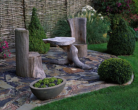 WOODEN_SEATS_AND_TABLE_ON_SLATE_TERRACE_WITH_TOPIARY_BOX_SHAPES_AND_WILLOW_FENCING_DESIGNER_JOHN_MAS