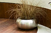 SPUN STAINLESS STEEL CONTAINER ON WOODEN DECK PLANTED WITH CAREX BUCHANANII. CONTAINER DESIGNED BY MARK PEDRO DE LA TORRE