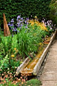 WATER FEATURE: A RILL RUNS ALONGSIDE A PATH WITH IRISES  PRIMULAS AND GEUMS.IN THE BACKGROUND IS CHARRED SPIRES  A TIMBER SCULPTURE BY ALSION CROWTHER. WINGWELL NURSERY   RUTLAND