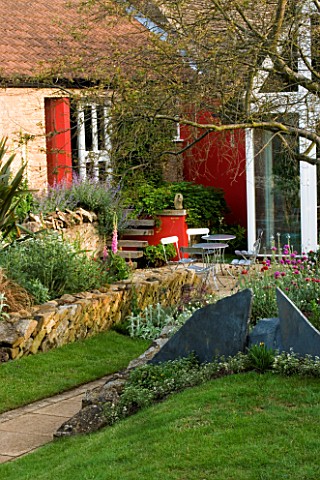 VIEW_ACROSS_THE_LAWN_TO_THE_PATIO_WITH_METAL_TABLE_AND_CHAIRS__RED_WALL_AND_ANGLED_DRYSTONE_WALL_WIN
