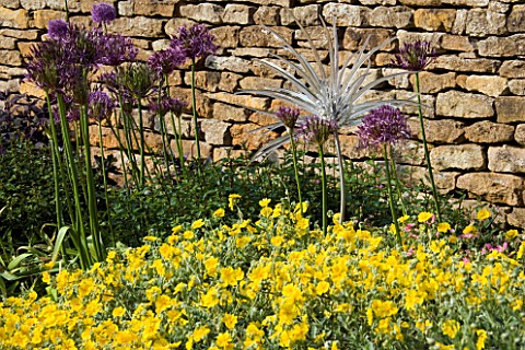 A_DELICATE_GLASS_SCULPTURE_BY_NEIL_WILKINS_IN_A_BORDER_BESIDE_A_STONE_WALL_WITH_ALLIUMS_AND_HELIANTH