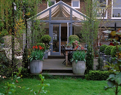LISETTE_PLEASANCE_GARDEN__LONDON_VIEW_TO_DECKED_TERRACE_AND_CONSERVATORY_WITH_LEAD_TABLE__CHAIRS__AN