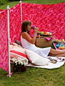 DESIGNER CLARE MATTHEWS: WIND BREAK SCREEN PROJECT - CLARE SITS IN THE WIND BREAK IN LAWN WITH CUSHIONS  BARBEQUE AND TRAY WITH DRINKS