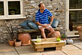 DESIGNER: CLARE MATTHEWS - LOW SLEEPER TABLE PROJECT: MAN WITH BLUE SHIRT SITS ON SEAT WITH LOW SLEEPER TABLE IN FRONT OF HIM ON PATIO
