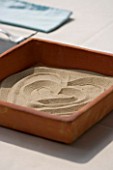DESIGNER: CLARE MATTHEWS - ZEN TRAY: SQUARE TERRACOTTA DISH ON TABLE WITH SAND MADE INTO A HEART SHAPE
